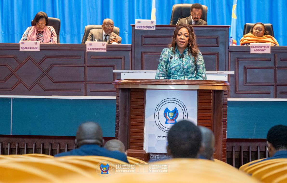 La députée Dorothée Madiya Mwamba, en veste à motifs verts, s'exprime à la tribune de l'Assemblée nationale de la RDC, derrière un pupitre portant l'emblème de l'institution ; les membres du présidium sont assis en surplomb.
