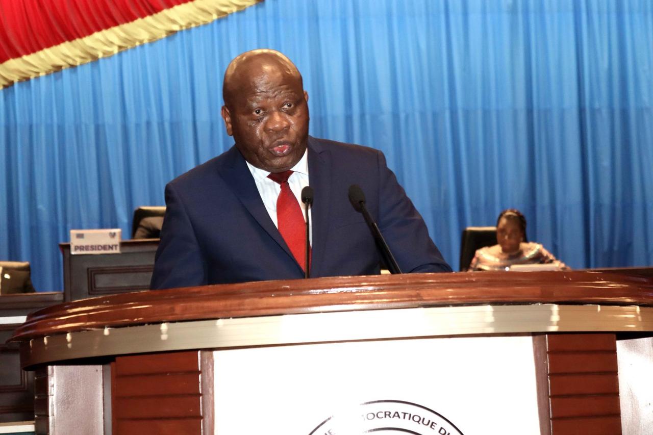 Un homme en costume bleu marine et cravate rouge parle dans deux micros, debout à la tribune en bois de l'Assemblée nationale congolaise, devant une tenture bleue et le drapeau de la RDC.