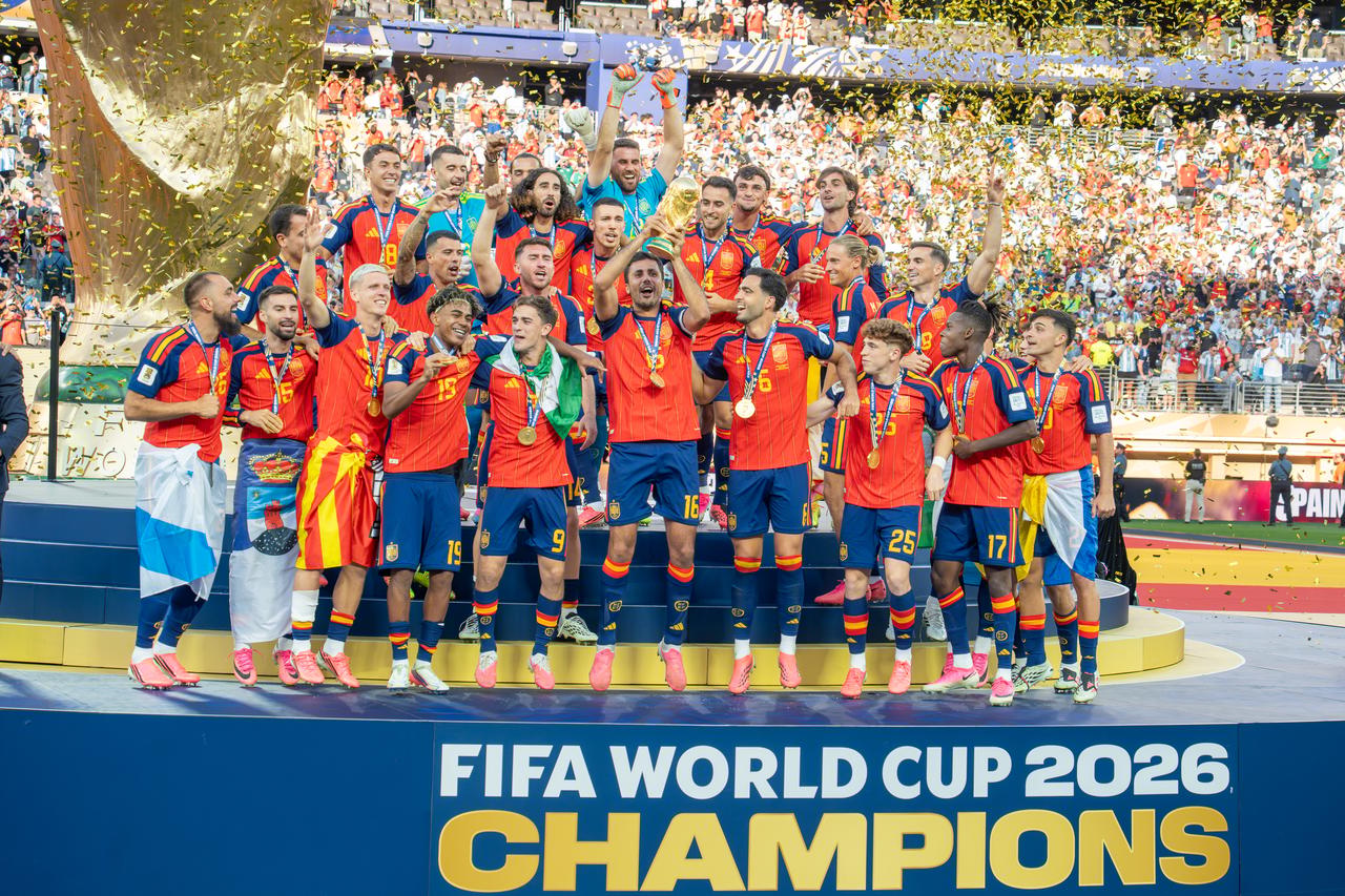 Les joueurs espagnols réunis sur le podium soulèvent le trophée de la Coupe du monde devant un panneau « FIFA World Cup 2026 Champions », sous les confettis.