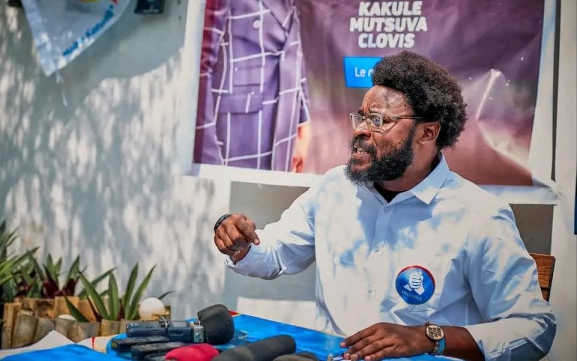 Clovis Mutsuva, en chemise bleu clair portant un badge du parti Ensemble pour la République, s'exprime devant plusieurs micros de presse, devant une banderole à son nom.