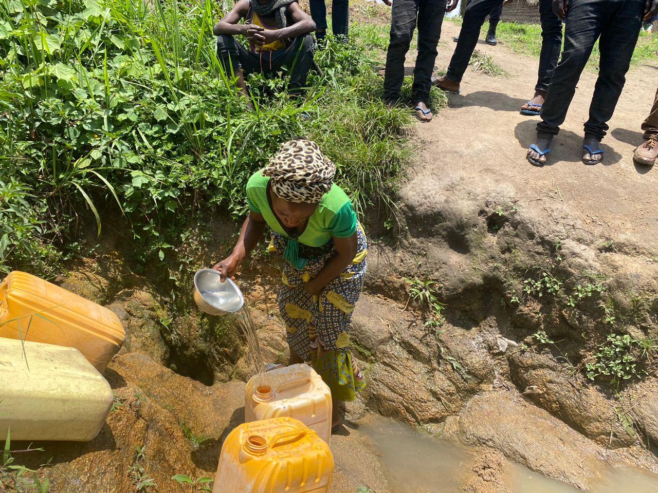 Une femme accroupie remplit des bidons jaunes avec une cuvette en puisant l'eau d'une source non aménagée creusée à même la terre, entourée de personnes debout au bord du trou.