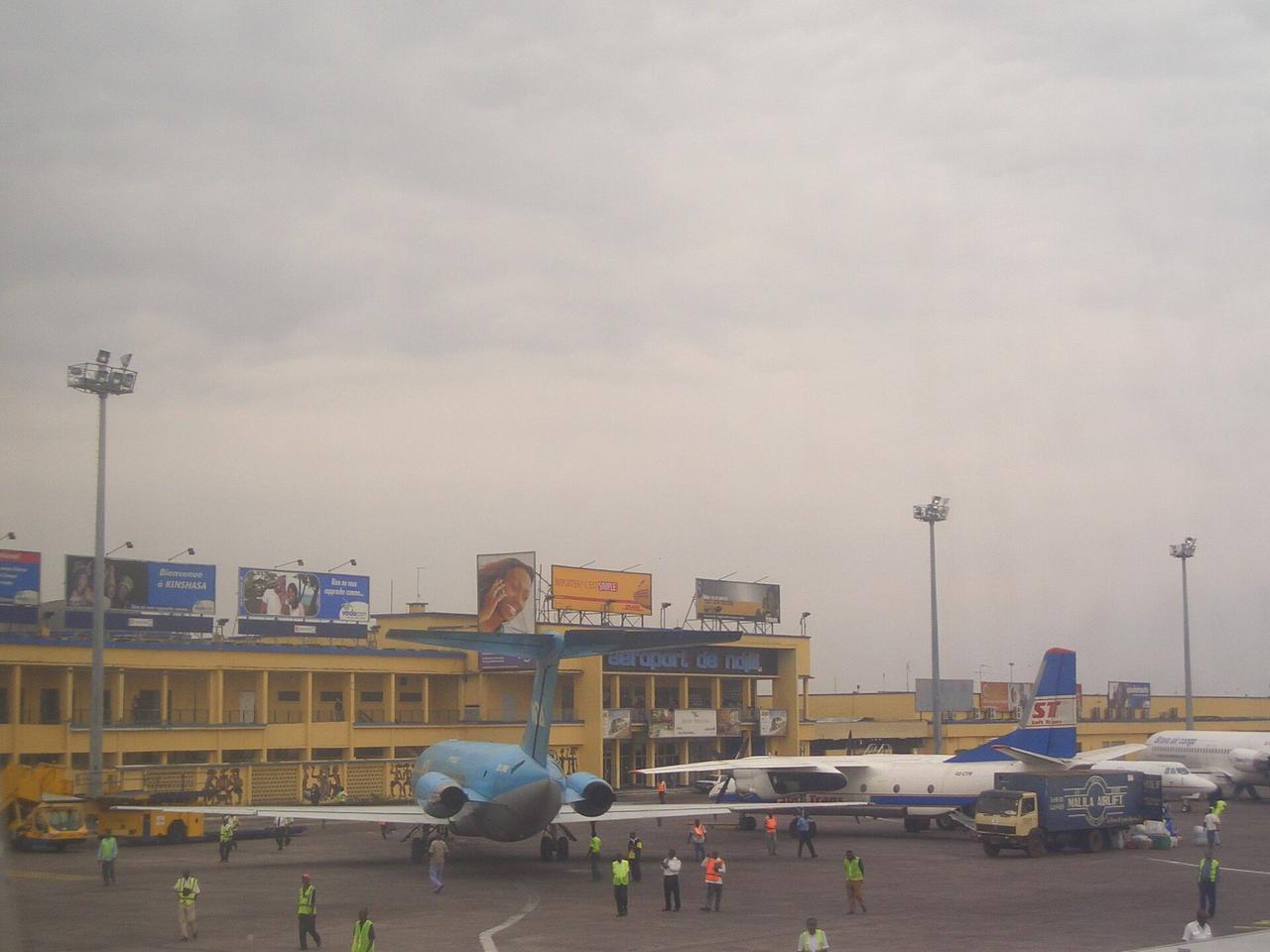 L'aérogare de l'aéroport international de N'Djili à Kinshasa, un long bâtiment jaune portant l'inscription « aéroport de ndjili » sur sa façade, avec plusieurs avions et des agents de piste stationnés sur le tarmac au premier plan.