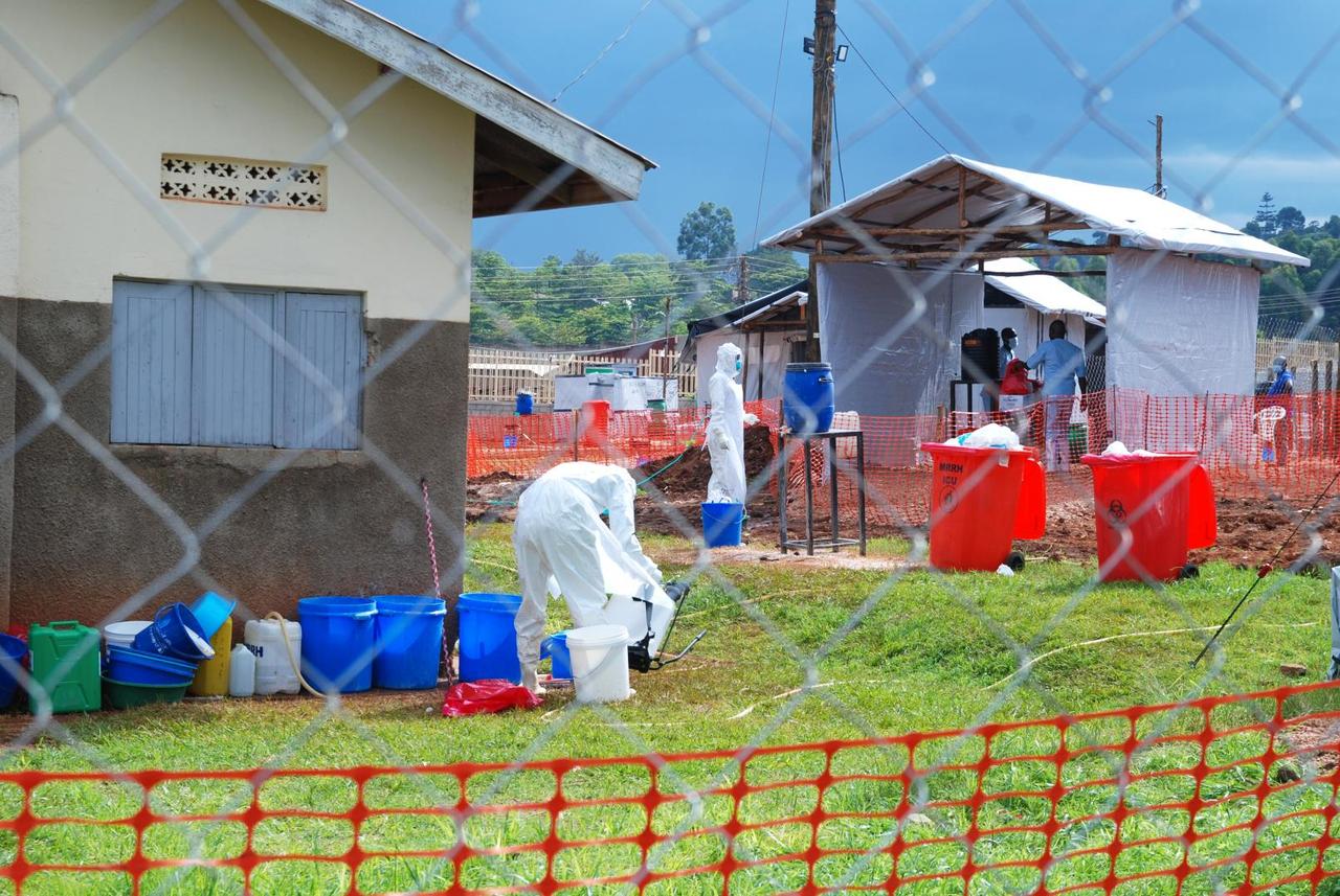 Agents de santé ougandais en combinaison de protection intégrale blanche au centre de traitement Ebola de l'hôpital régional de Mubende : l'un désinfecte du matériel au-dessus de seaux, tentes d'isolement bâchées et poubelles rouges à déchets biologiques à l'arrière-plan, derrière une clôture grillagée et un filet de sécurité orange.