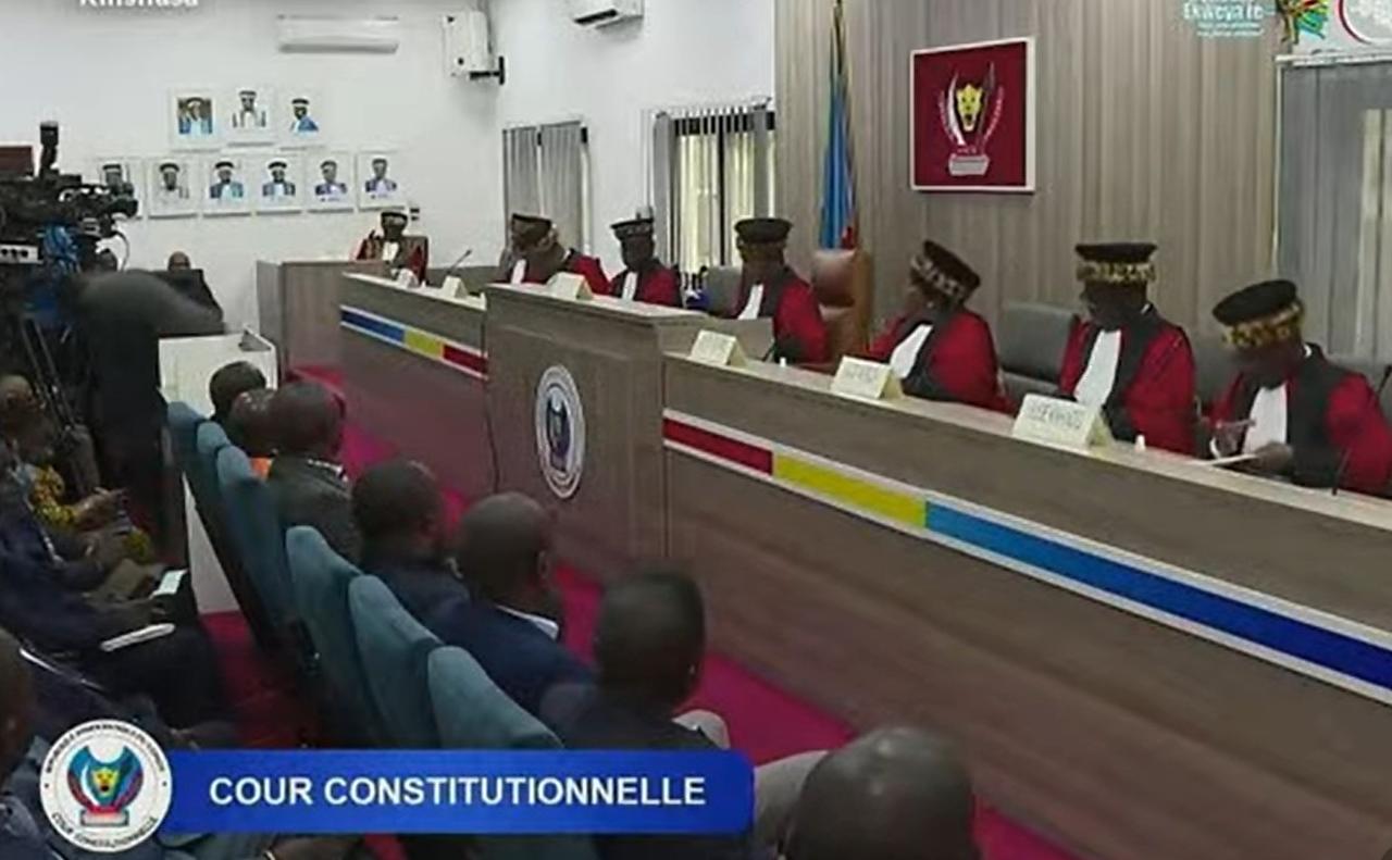 Les juges de la Cour constitutionnelle de la RDC, en toge rouge et toque bordée de peau de léopard, siègent en audience publique à Kinshasa, devant les armoiries de la République ; au premier plan, l'assistance et les caméras.