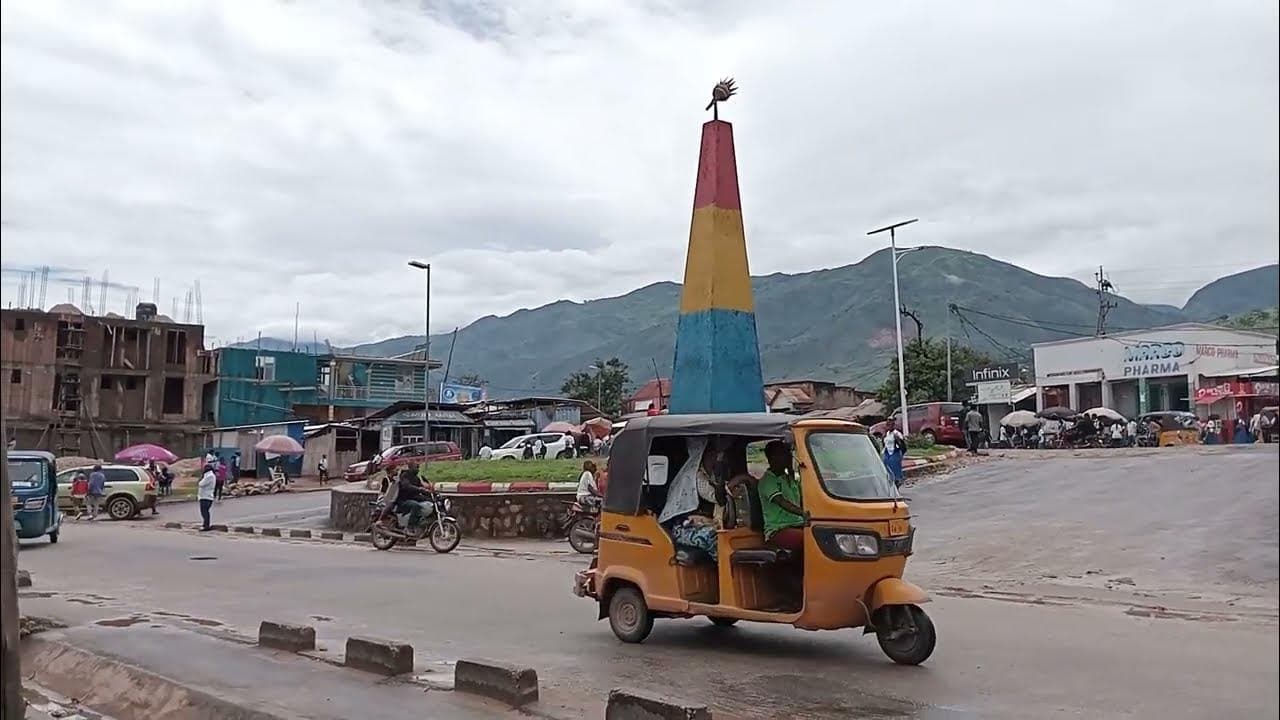 Scène de rue au centre-ville d'Uvira : un bajaj jaune et des motos passent devant un rond-point orné d'un monument aux couleurs du drapeau congolais, bordé de boutiques, avec les montagnes en arrière-plan.