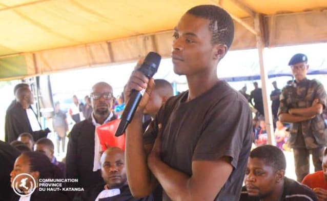 Un jeune homme debout, un micro à la main, répond aux questions du tribunal militaire de garnison de Lubumbashi lors d'une audience foraine sous une tente ; des avocats en robe noire et un militaire en béret bleu l'entourent.