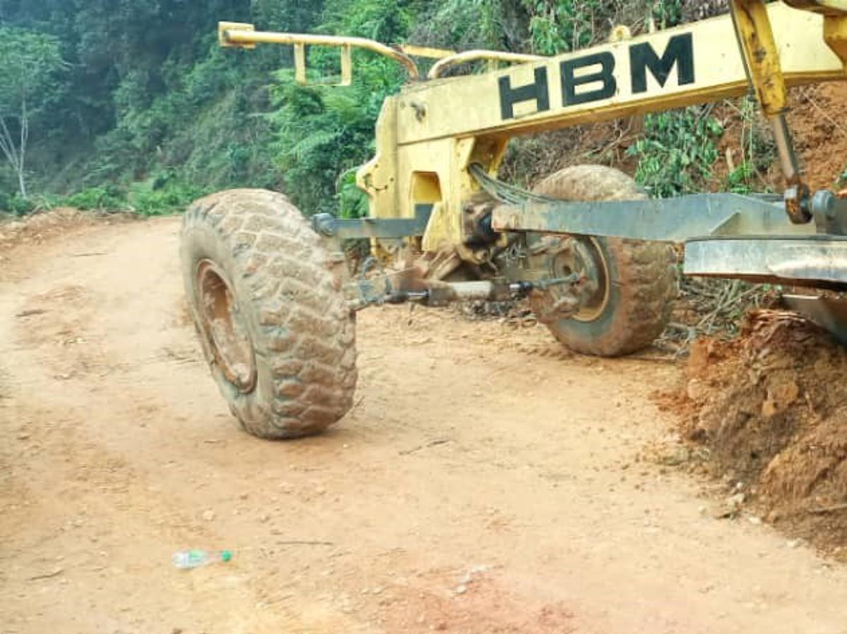 Une niveleuse jaune de marque HBM, aux gros pneus couverts de boue, travaille sur une route de terre fraîchement ouverte, taillée à flanc de colline à travers la forêt dense ; la terre ocre retournée s'entasse sur le bas-côté.