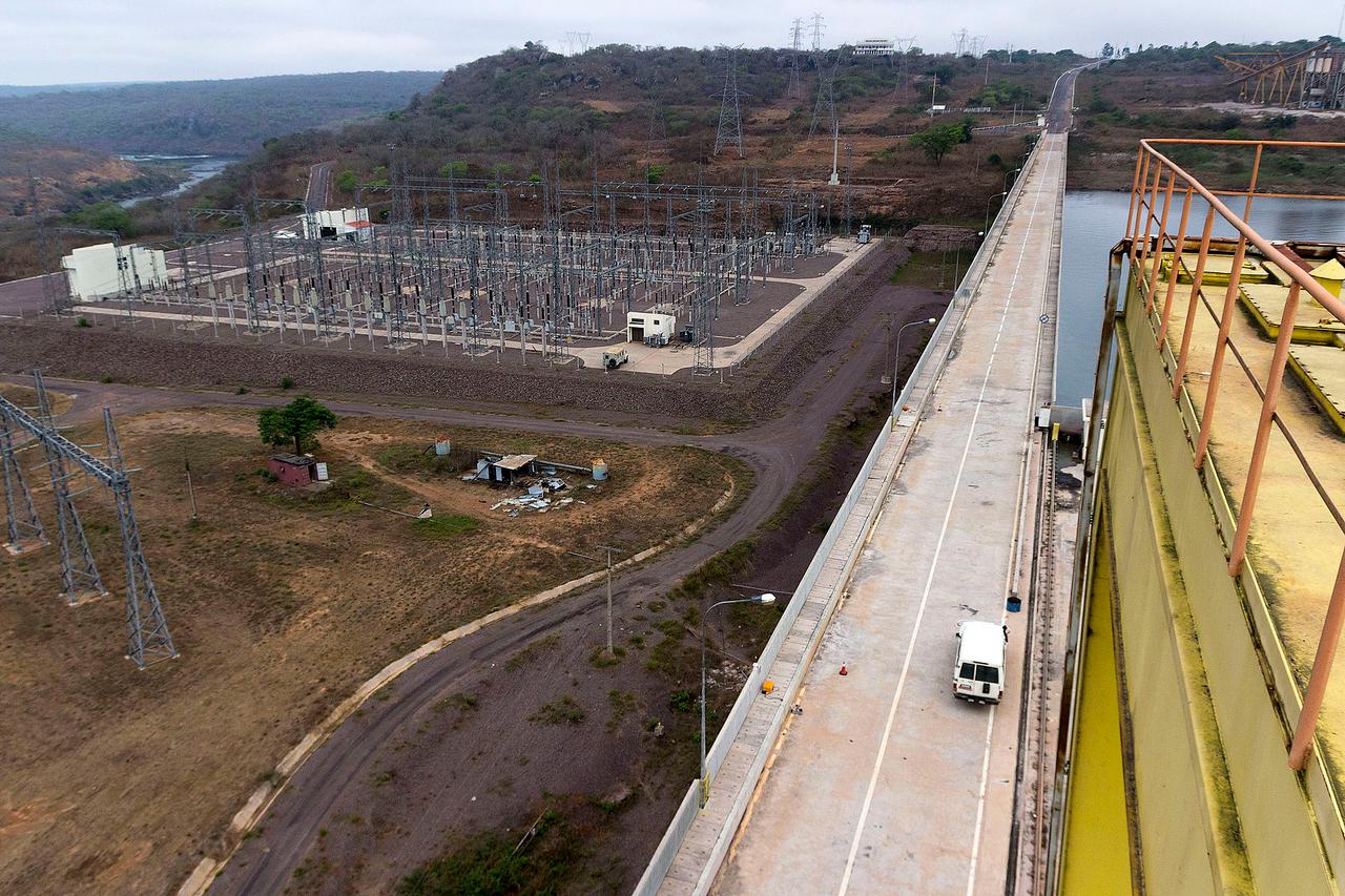 Vue aérienne d'un poste électrique à haute tension en Angola : rangées de portiques et d'appareillage métalliques, pylônes et lignes électriques s'éloignant vers des collines, le long de la crête d'un barrage hydroélectrique surplombant une retenue d'eau.