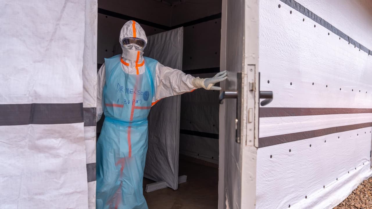 Un soignant en combinaison de protection intégrale, capuche, lunettes et gants, ouvre la porte d'une unité d'isolement Ebola aux parois de bâche blanche ; son nom, « Dr Nelson », et l'heure « 1.30 PM » sont inscrits au marqueur sur son tablier bleu.