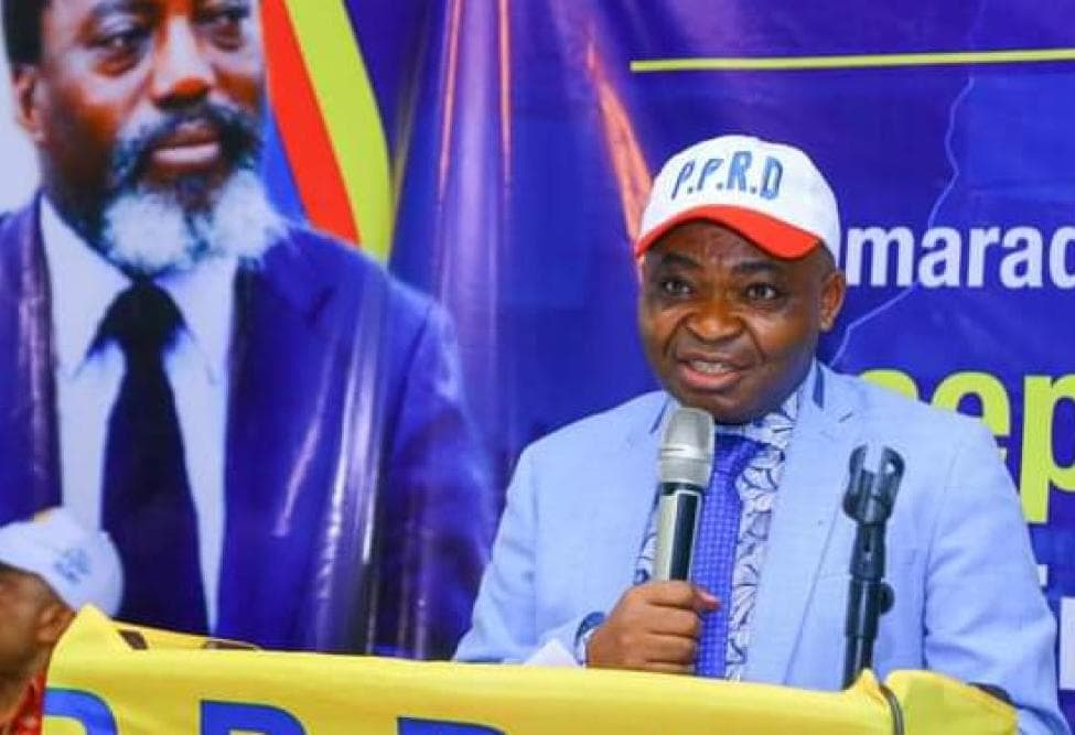 Ferdinand Kambere, secrétaire permanent adjoint du PPRD, parle au micro derrière un pupitre, coiffé d'une casquette blanche siglée P.P.R.D, devant une affiche à l'effigie de Joseph Kabila et une banderole du parti