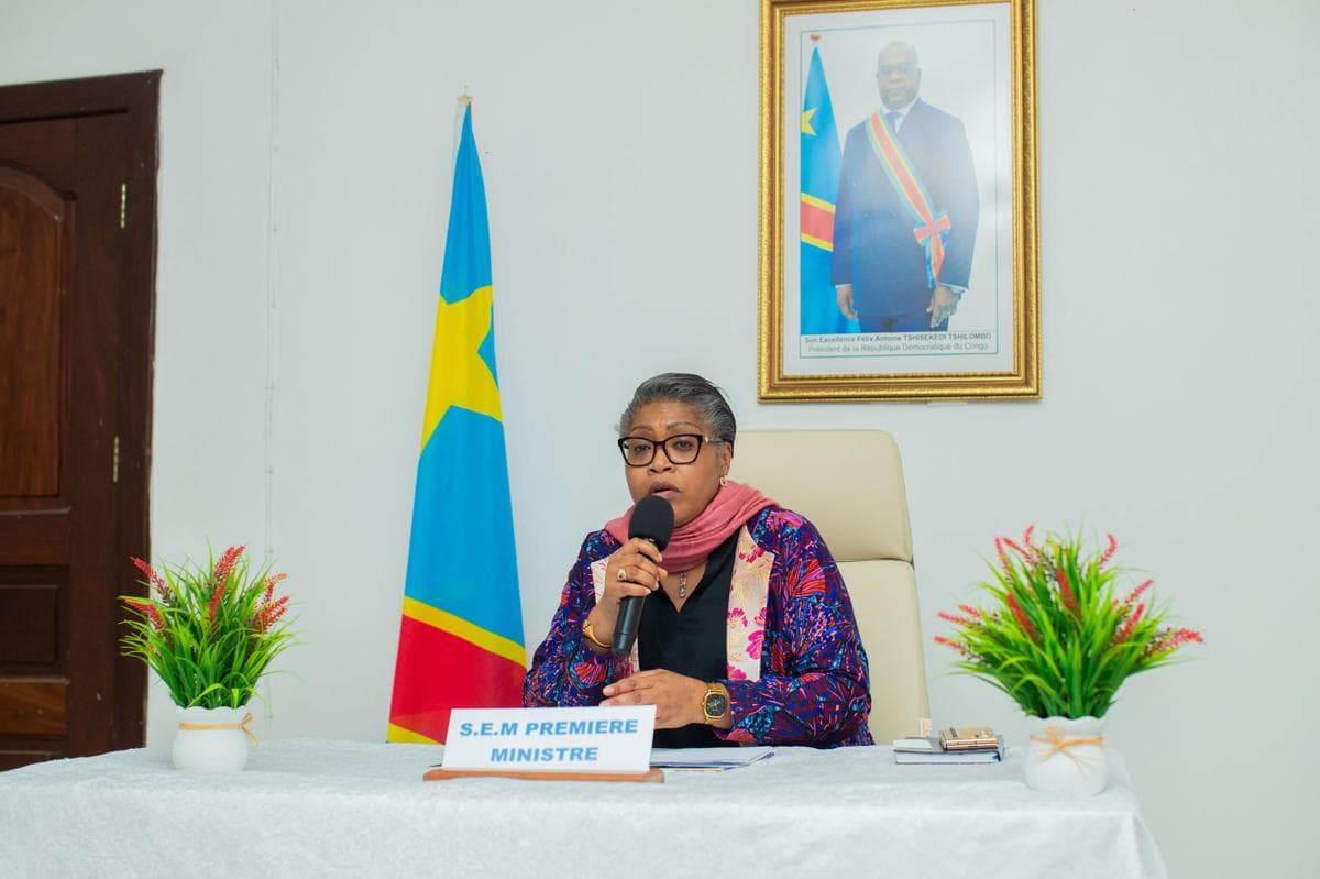 La Première ministre Judith Suminwa Tuluka s'exprime au micro derrière un pupitre « S.E.M Première ministre », devant le drapeau de la RDC et le portrait du président Tshisekedi.